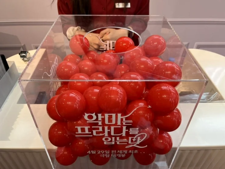 A transparent box filled with red balls, possibly for an interactive event, with a logo affixed to its side, adding an element of playful engagement. - The Devil Wears Prada 2: An Exclusive Pop-Up Showroom Experience in Seoul's Seongsu District