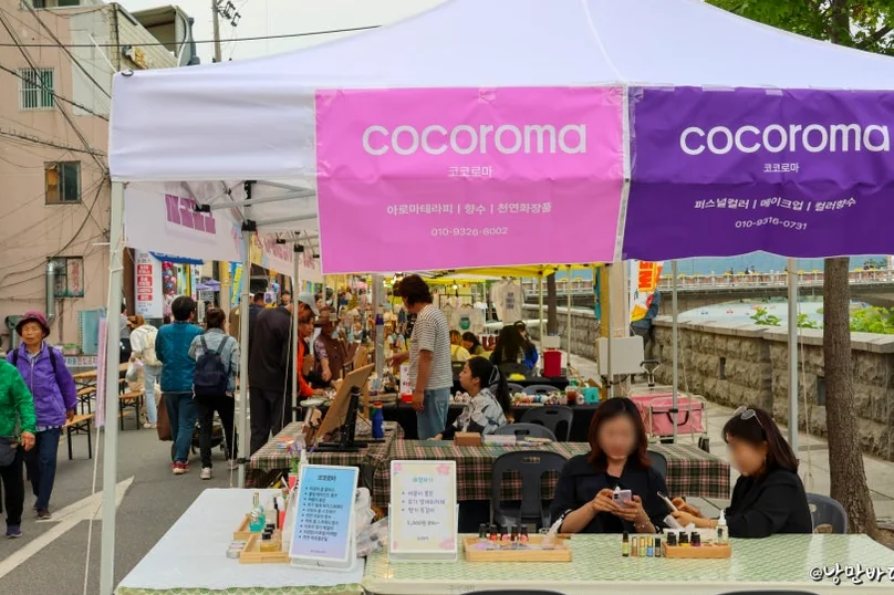 A bustling daytime market scene with various vendor tents, including one with 'cocoroma' signs, selling crafts and interacting with customers. - The 68th Miryang Arirang Festival: A Timeless Korean Cultural Spectacle