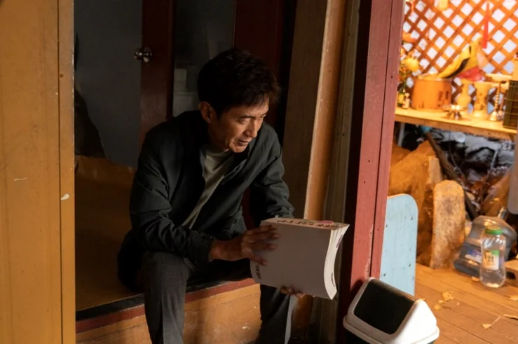 alt: 素朴な室内の設定で、物思いにふけるキャラクターを捉えた情緒的なスチールカット - 呪術と権力の狂騒曲、韓国映画『神明』が暴く2026年の真実と聖地巡礼の記録