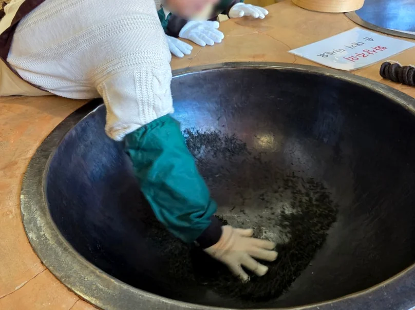 alt="博物館の体験センターで、大きな鉄釜を使って手作業で茶葉を焙煎する伝統的な「ドックム」工程のクローズアップ" - 智異山の静寂を味わう：河東（ハドン）野生茶博物館で体験する韓国最高級のお茶文化ガイド