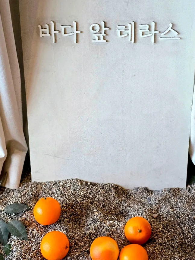 A sun-drenched rooftop terrace featuring a magnificent artificial orange tree bursting with fruit, surrounded by rustic baskets overflowing with fresh oranges, creating a vibrant and unique outdoor ambiance against a clear blue sky. - Badaap Terrace: Yeongjongdo's Premier Oceanfront Cafe Experience