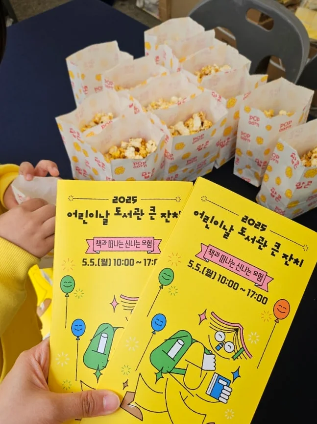 A young visitor holding colorful event pamphlets and popcorn snacks, illustrating the tactile and joyful experience of participating in the festivities. - Literary Luxury in Gangnam: The 2026 Great Children’s Day Festival at the National Library for Children and Young Adults