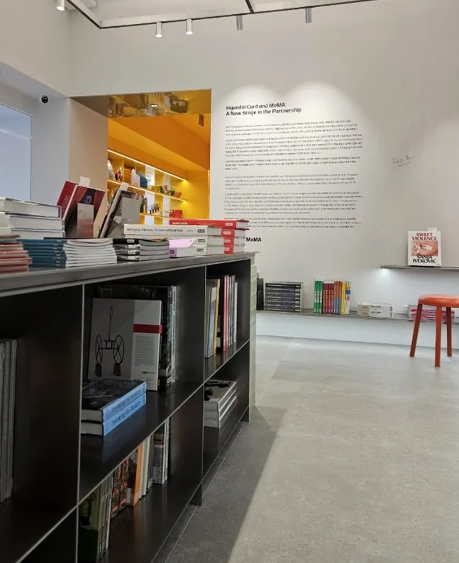 A detailed interior view of the MoMA Bookstore, showcasing a corner with an iconic orange design stool amidst neatly organized books and a subtle descriptive text panel on the wall, inviting a moment of thoughtful pause.</div - MoMA Bookstore Seoul: A Curated Sanctuary for Art and Design