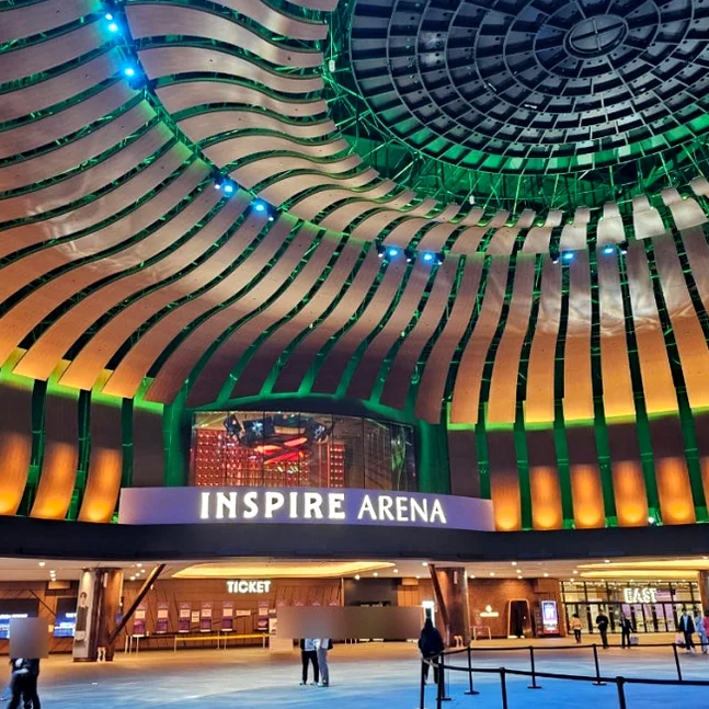 The grand architectural entrance of the Inspire Arena, showcasing a sophisticated blend of wood-toned curves and emerald-green lighting that anchors the resort's identity. - Neon Gastronomy: The Inspire Aurora Night Market Lifestyle Guide