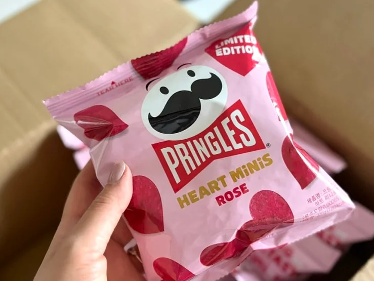開封されたプリングルズ・ハート・ミニ・ローズの箱に手が伸び、ピンクを基調としたパッケージを一つ取り出す様子。アンボクシング（開封）の触覚的な喜びと発見を強調。 - 韓国限定・世界初の上陸。メガボックスとGSザ・フレッシュで出会う「プリングルズ・ハート・ミニ・ローズ」の誘惑