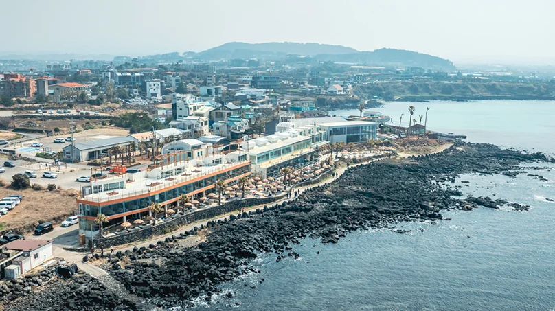 An aerial view of the coastal cafe building complex and surrounding rocky shoreline. - 済州島・涯月（エウォル）の海を飲み込む贅沢。韓屋と絶景が織りなす『ヘジゲ（海知開）』完全ガイド
