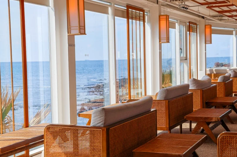 A cafe interior with large windows showcasing an ocean view, and empty wooden and rattan seating. - 済州島・涯月（エウォル）の海を飲み込む贅沢。韓屋と絶景が織りなす『ヘジゲ（海知開）』完全ガイド