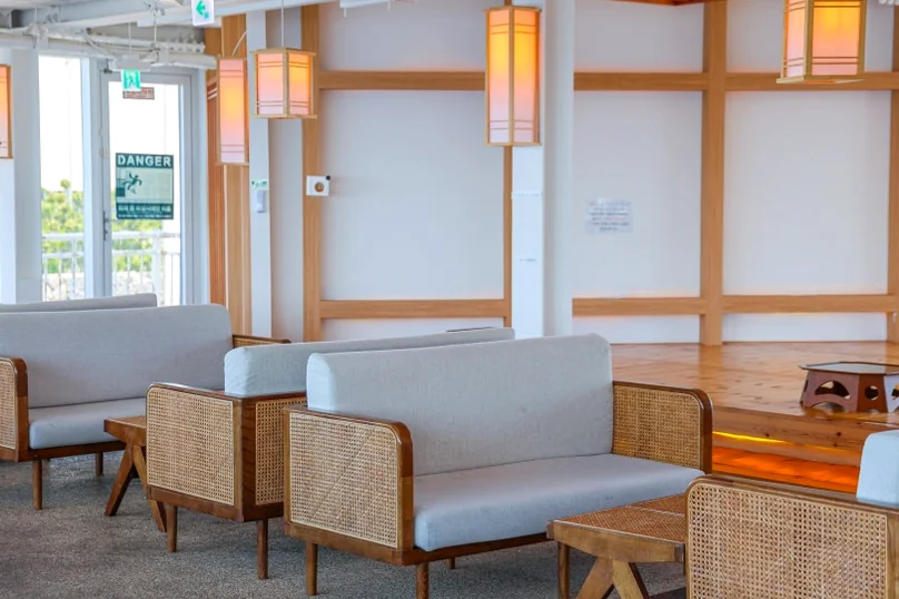 A long cafe interior featuring traditional Korean lanterns, wooden lattice screens, and patrons seated by the windows. - 済州島・涯月（エウォル）の海を飲み込む贅沢。韓屋と絶景が織りなす『ヘジゲ（海知開）』完全ガイド