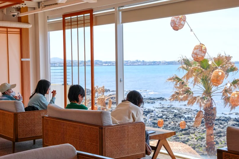Cafe patrons seated in rattan chairs, looking out large windows at the ocean and a palm tree decorated with lanterns. - 済州島・涯月（エウォル）の海を飲み込む贅沢。韓屋と絶景が織りなす『ヘジゲ（海知開）』完全ガイド