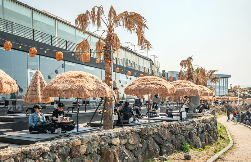 An outdoor seating area with straw umbrellas and tables, located on a stone wall overlooking the ocean. - 済州島・涯月（エウォル）の海を飲み込む贅沢。韓屋と絶景が織りなす『ヘジゲ（海知開）』完全ガイド