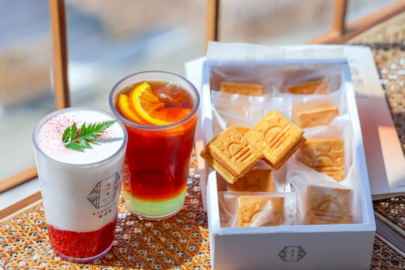 Two glasses of beverages, one with orange slices and another with foam, next to a white box of Jeju-style pastries on a patterned table. - 済州島・涯月（エウォル）の海を飲み込む贅沢。韓屋と絶景が織りなす『ヘジゲ（海知開）』完全ガイド