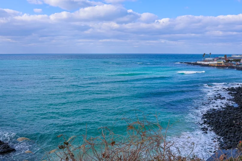A wide view of the turquoise ocean with waves breaking on a rocky shore, under a partly cloudy sky. - 済州島・涯月（エウォル）の海を飲み込む贅沢。韓屋と絶景が織りなす『ヘジゲ（海知開）』完全ガイド