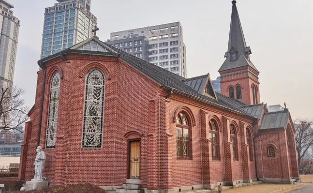 Frontal exterior view of Yakhyun Catholic Church, a red brick building with a prominent spire, under a bright sky with sun flare. - ソウル駅の裏側に眠る130年の物語。中林洞・薬峴聖堂(ヤッキョンソンダン)で巡る歴史と美学の散歩道