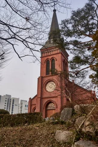 Vertical view of the main facade of Yakhyun Catholic Church, showcasing a large wooden double door, a circular window, and a tall spire. - ソウル駅の裏側に眠る130年の物語。中林洞・薬峴聖堂(ヤッキョンソンダン)で巡る歴史と美学の散歩道