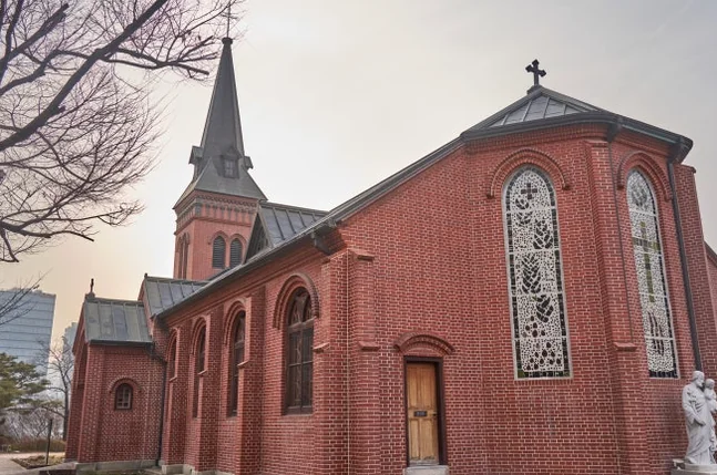 Exterior of Yakhyun Catholic Church, partially framed by a large bare tree, with soft sunlight illuminating the side of the building. - ソウル駅の裏側に眠る130年の物語。中林洞・薬峴聖堂(ヤッキョンソンダン)で巡る歴史と美学の散歩道