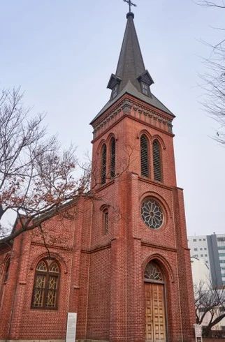 Low-angle perspective of Yakhyun Catholic Church's spire, surrounded by bare tree branches and natural ground textures. - ソウル駅の裏側に眠る130年の物語。中林洞・薬峴聖堂(ヤッキョンソンダン)で巡る歴史と美学の散歩道
