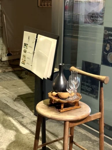 A wooden stool with a ceramic bottle and glasses next to a menu stand - 2026年ソウル・新堂洞の最旬スポット。伝統酒ソムリエが贈る至福の韓食ダイニング『ピョジュ(表酒)』へ。