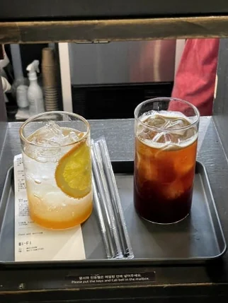 A glass of orange ade and a glass of iced coffee on a metal tray in a delivery slot - 郵便局がカフェに？ソウル・新堂洞(シンダンドン)の隠れ家『メールルーム』で体験する非日常