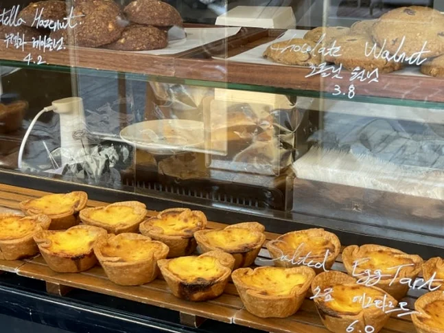 Rows of egg tarts and cookies on wooden trays inside a glass display case - 郵便局がカフェに？ソウル・新堂洞(シンダンドン)の隠れ家『メールルーム』で体験する非日常