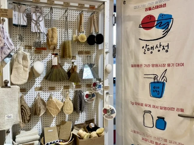 Eco-friendly cleaning tools on a pegboard next to a fabric banner with the store logo - 2026年最新｜ソウル・望遠洞(マンウォンドン)のエコ＆感性散策。ゼロウェイストの聖地'アルメン商店'と南仏カフェ'リトルメドウ'