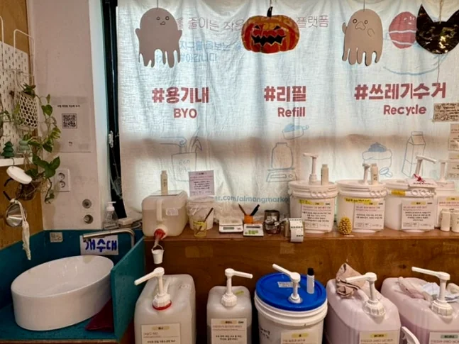 Refill station area with bulk liquid containers and recycling slogans on a white curtain - 2026年最新｜ソウル・望遠洞(マンウォンドン)のエコ＆感性散策。ゼロウェイストの聖地'アルメン商店'と南仏カフェ'リトルメドウ'