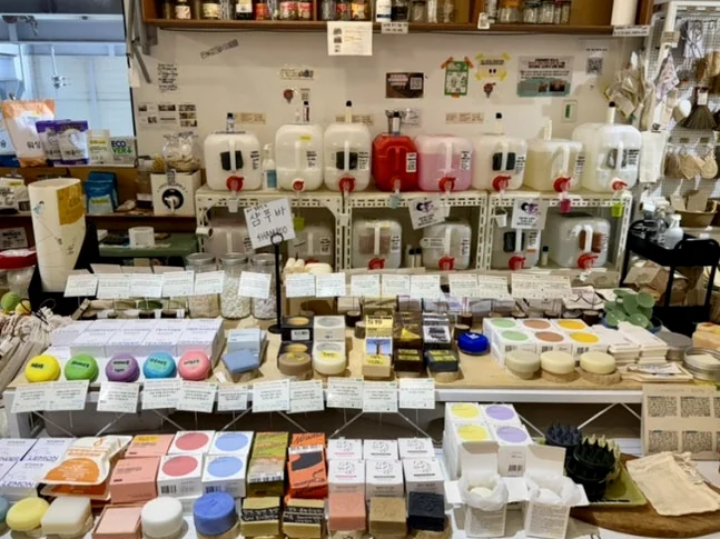 Organized display of colorful solid soaps and bulk detergent containers on a white table - 2026年最新｜ソウル・望遠洞(マンウォンドン)のエコ＆感性散策。ゼロウェイストの聖地'アルメン商店'と南仏カフェ'リトルメドウ'
