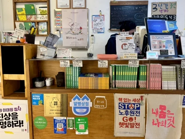 Wooden checkout counter featuring various social campaign stickers and informational pamphlets - 2026年最新｜ソウル・望遠洞(マンウォンドン)のエコ＆感性散策。ゼロウェイストの聖地'アルメン商店'と南仏カフェ'リトルメドウ'