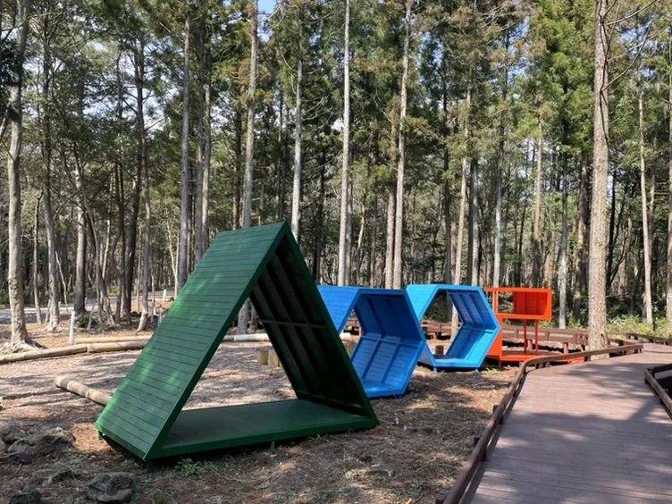Three colorful abstract seating structures, green, blue, and orange, positioned along a wooden boardwalk in a forest. - 済州島、心癒される森の道へ：水岳オルムとモチェワッの秘密 🌿