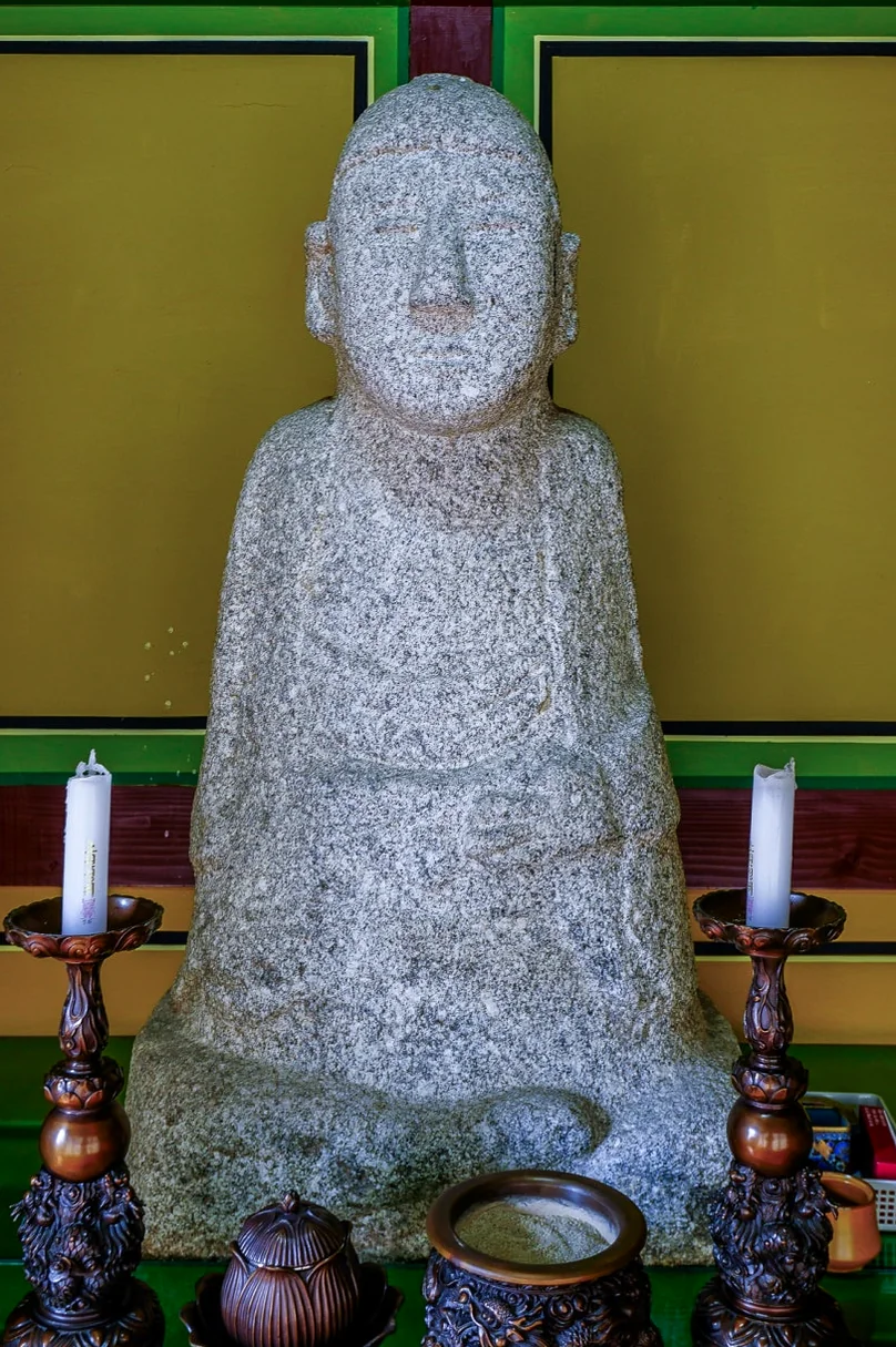 A grey stone Buddha statue seated inside a temple hall, flanked by two candle holders and incense burners on a table. - 全州(チョンジュ)西古寺(ソゴサ)で心癒すテンプルステイ体験：静寂の中で見つける本当の自分