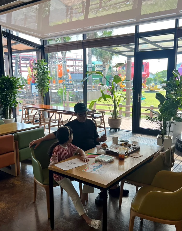 Wooden table and chairs in a cafe with large glass windows and indoor plants - 済州島・涯月(エウォル)の静かな休息地。ミッカン倉庫(Mikkang Changgo)で過ごす、蜜柑の香りに包まれた休日