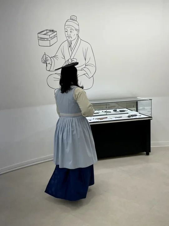 한복을 곱게 차려입은 방문객의 뒷모습. 전시된 유물을 감상하는 모습에서 문화적인 분위기와 자연스러운 연출이 느껴집니다. - ソウル薬令市韓医学博物館：五感で巡る、韓国伝統医療の魅力と癒し体験🌿
