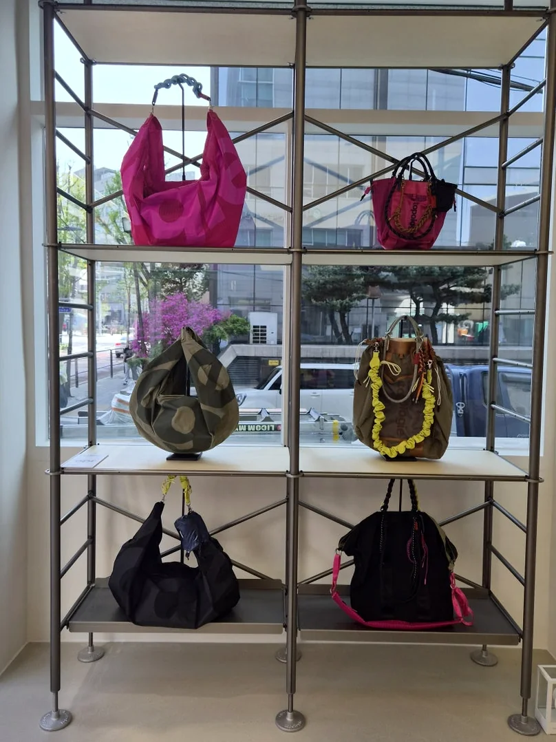 Close-up view of colorful Topologie bags displayed on a metal shelf near a glass entrance - ソウルの森に誕生した Topologie 韓国初フラッグシップ：2026 SS ‘The Starbound’ の世界 🧭
