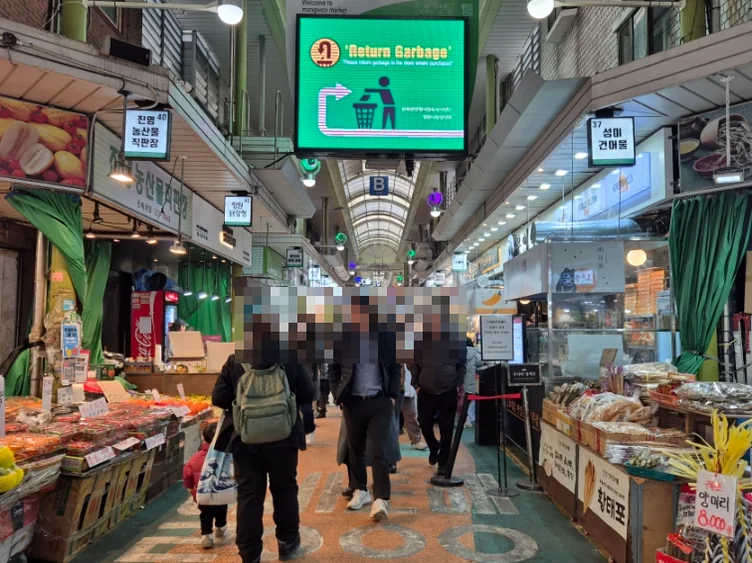 A long market aisle with diverse stalls, a large green display screen, and blurred people. - Q's Dakgangjeong: A Culinary Gem in Seoul's Bustling Mangwon Market