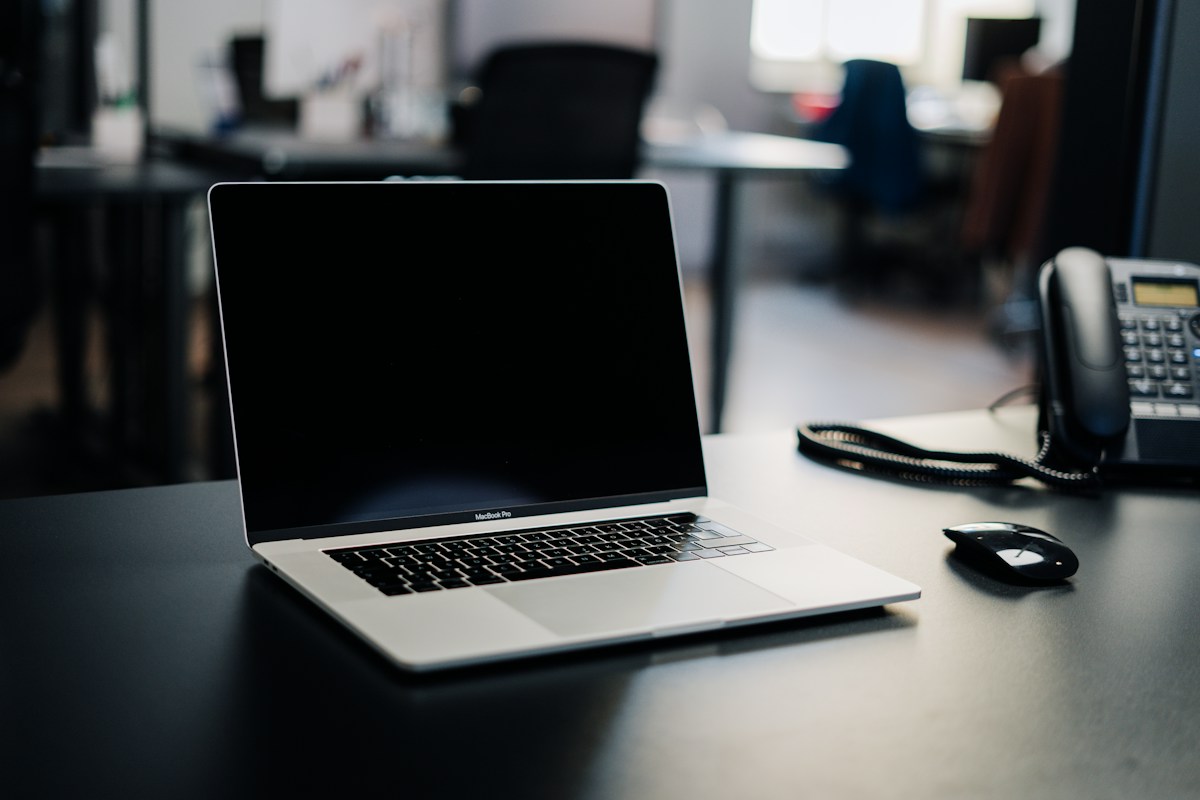 MacBook Pro laptop with a dark screen on a black office desk, next to a black mouse and a desk phone