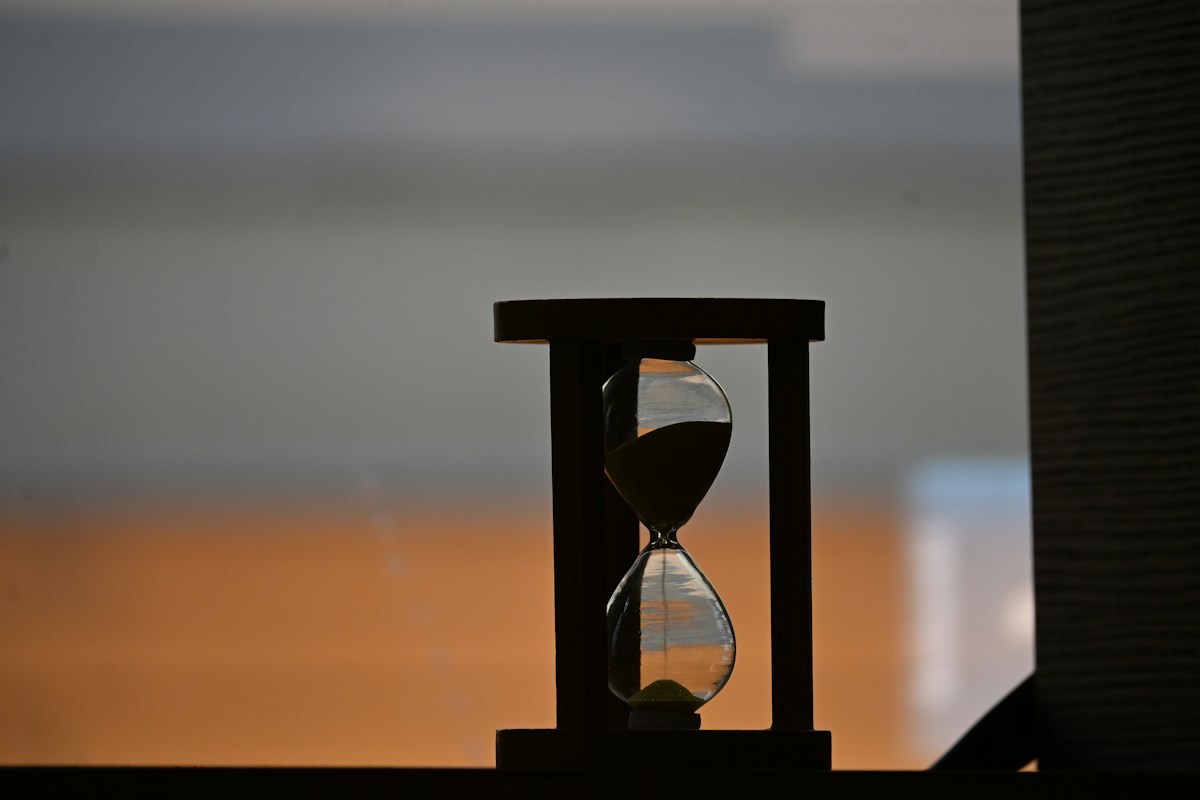 Silhouette of a wooden-framed hourglass, sand draining from the top bulb, against a blurred orange and grey background