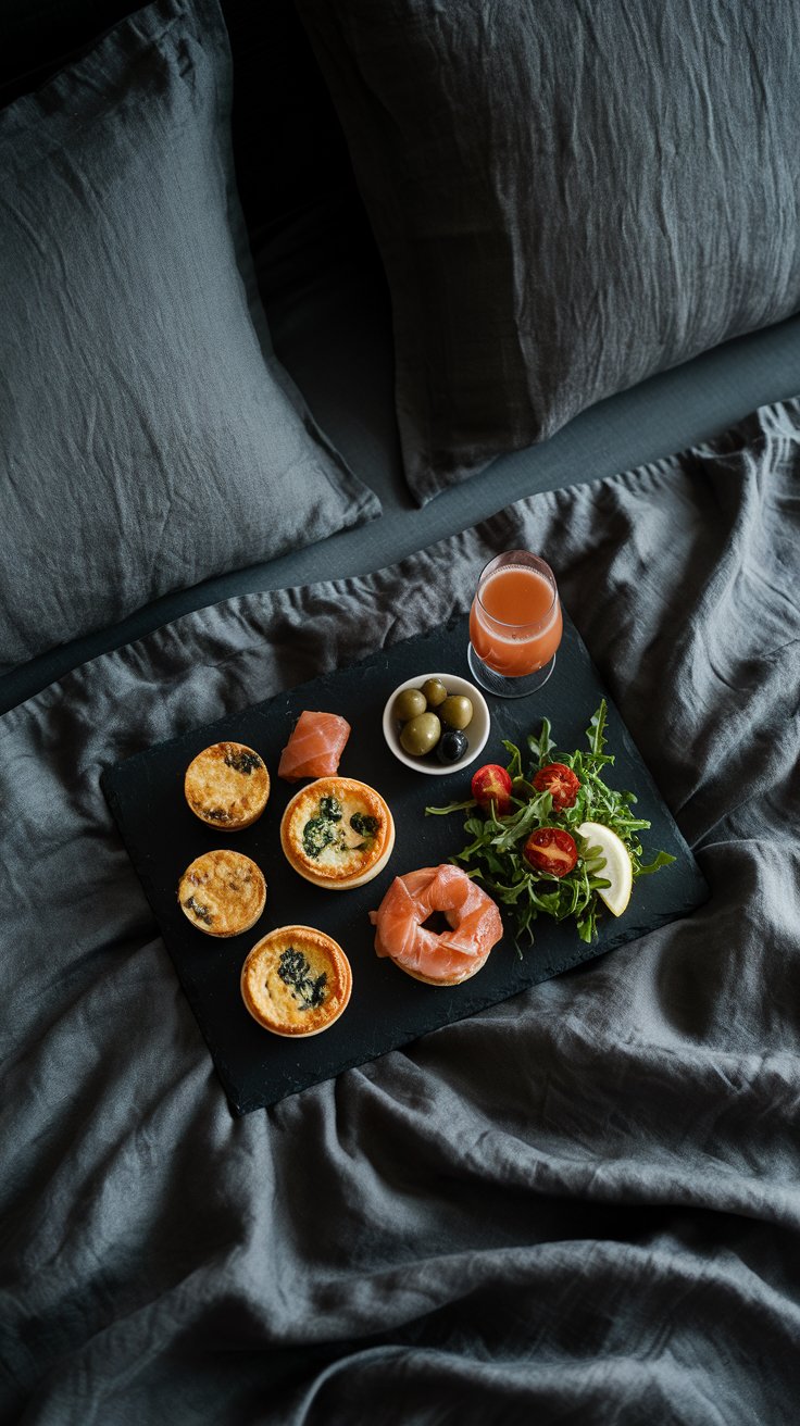 luxurious breakfast in bed, savory brunch style, dark grey linen bedding, slate tray, mini quiches (lorraine or spinach and feta), smoked salmon and cream cheese bagels (mini bagels), arugula salad with lemon vinaigrette, cherry tomatoes, small dish of olives, glass of Bloody Mary or Mimosa