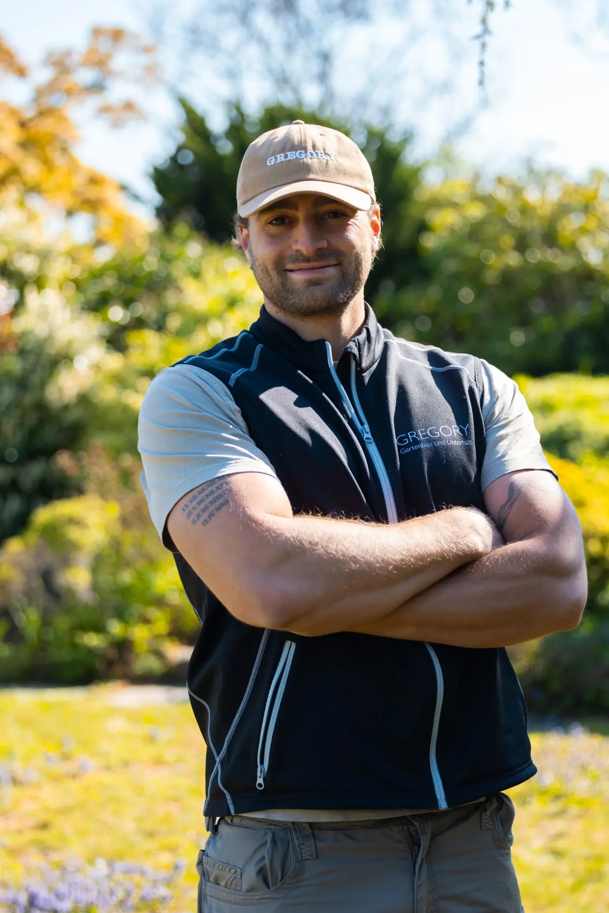 Gregory Wurmser, lächelnd, Arme verschränkt, GREGORY-Gilet und Cap im Garten