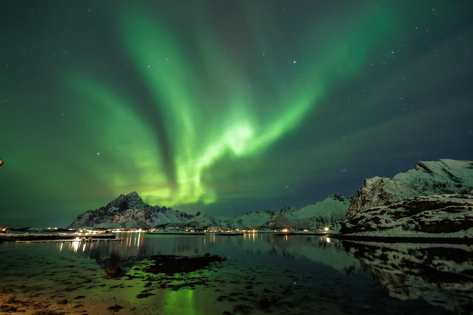 Green aurora curtains over snowy mountains and a still fjord in Norway.
