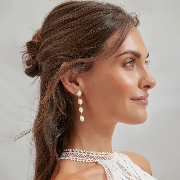 Une femme aux cheveux bruns mi-longs, coiffés en demi-attaché, elle porte des boucles d’oreilles pendantes dorées avec des perles.