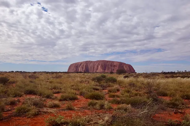 Vue panoramique d'Uluru (Ayers Rock), le monolithe de grès rouge emblématique, s'élevant au-dessus de la plaine désertique australienne sous un ciel nuageux.